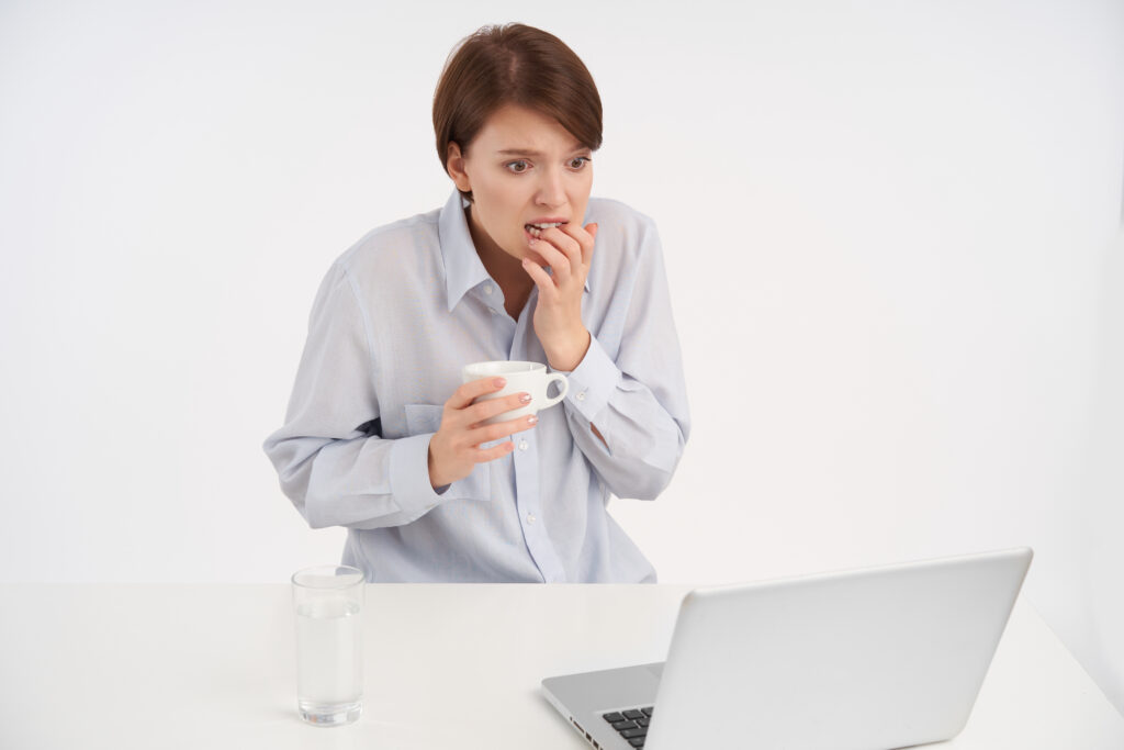 Shocked young pretty short haired brunette lady rounding her eyes while looking scaredly at screen of laptop and biting raised hand, sitting over white background in formal clothes